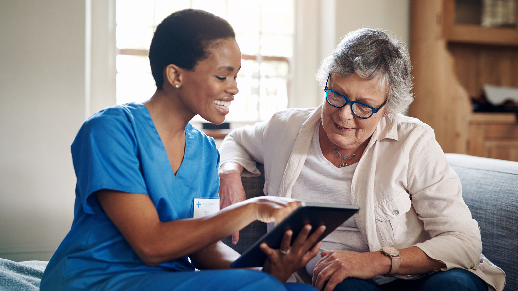  Senior woman using a digital tablet with a nurse on the sofa.