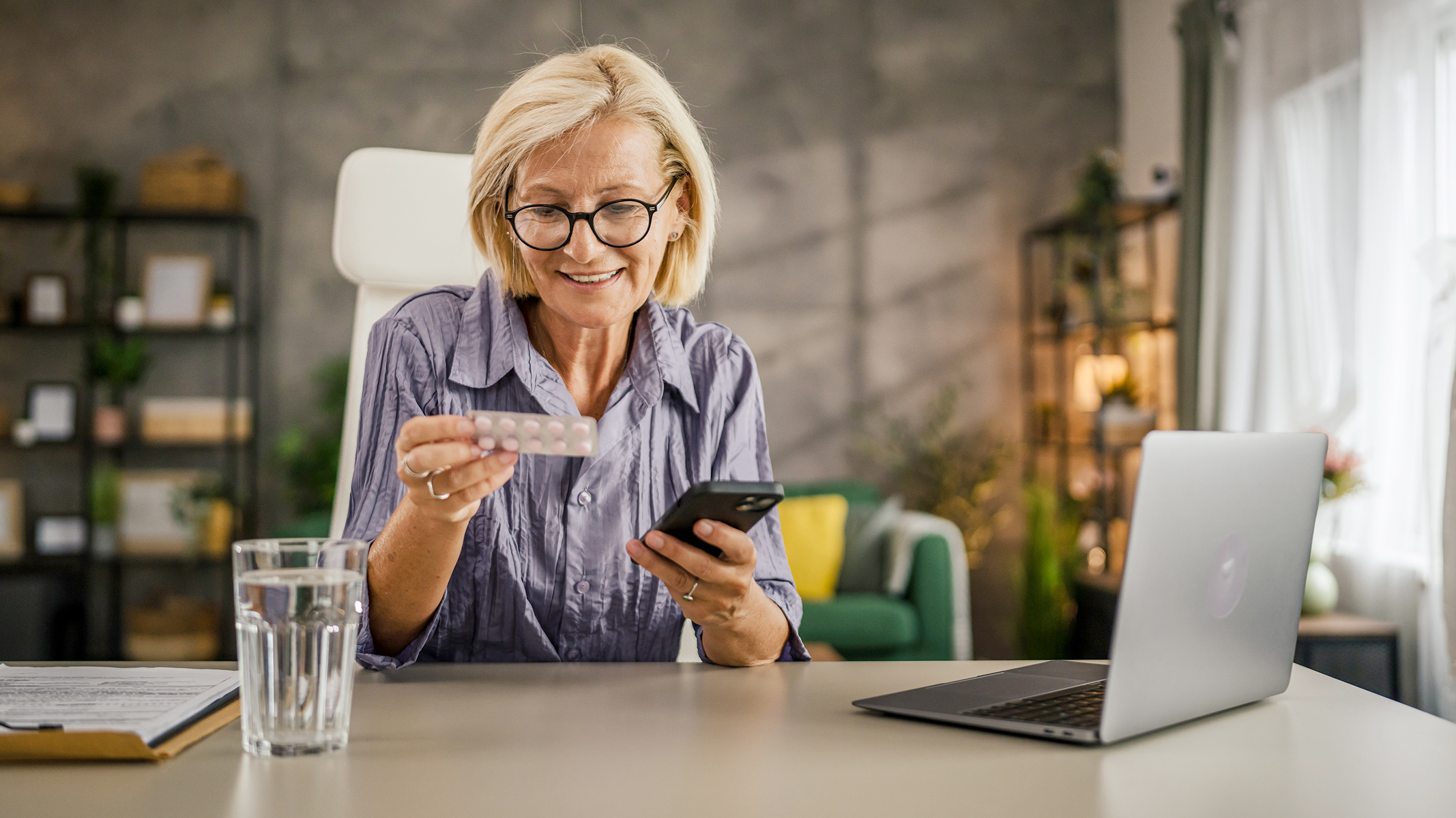 Woman looking at prescription medication with phone in hand and laptop nearby.