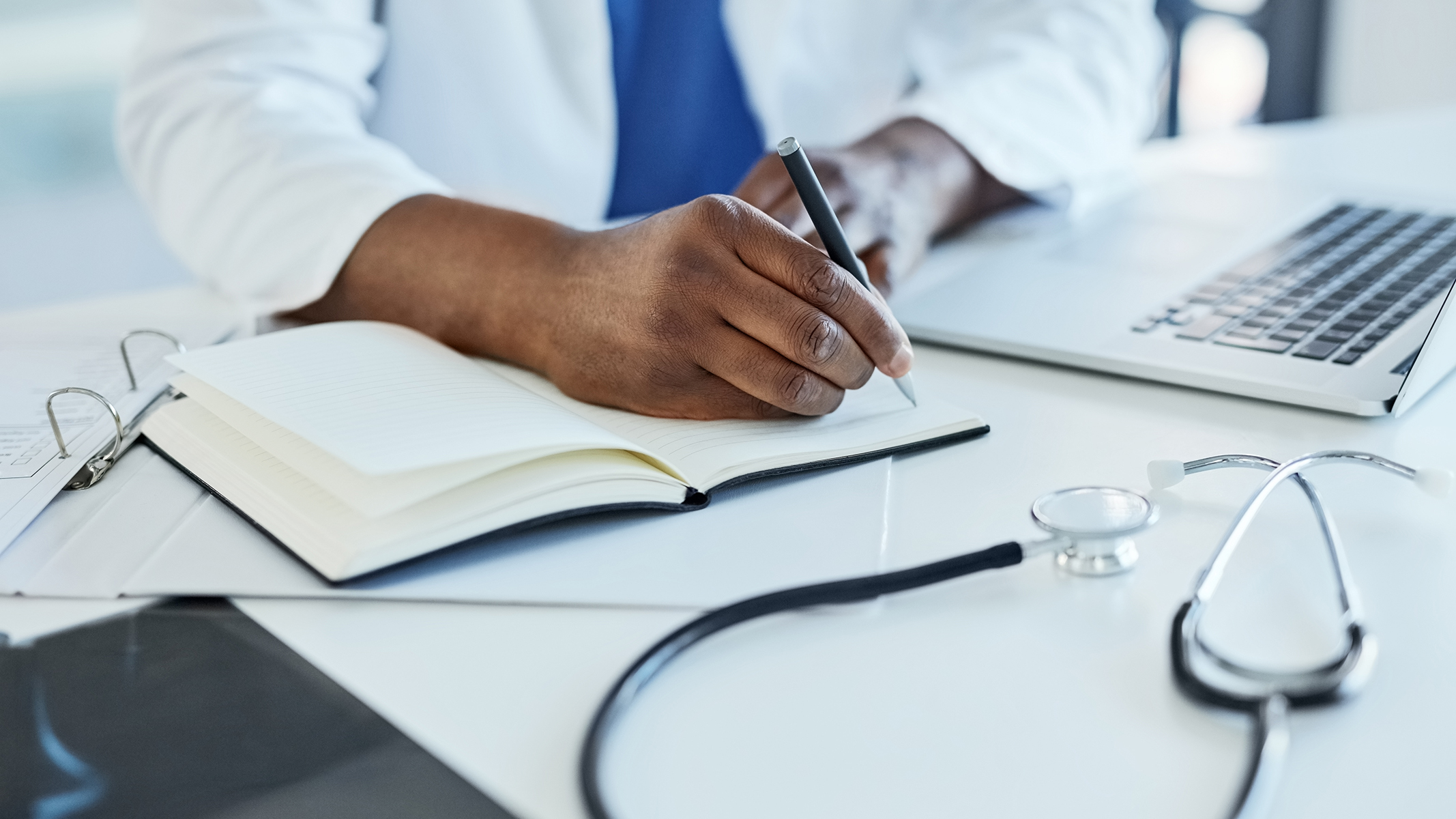 Primer plano de un médico usando un computador portátil y tomando notas.