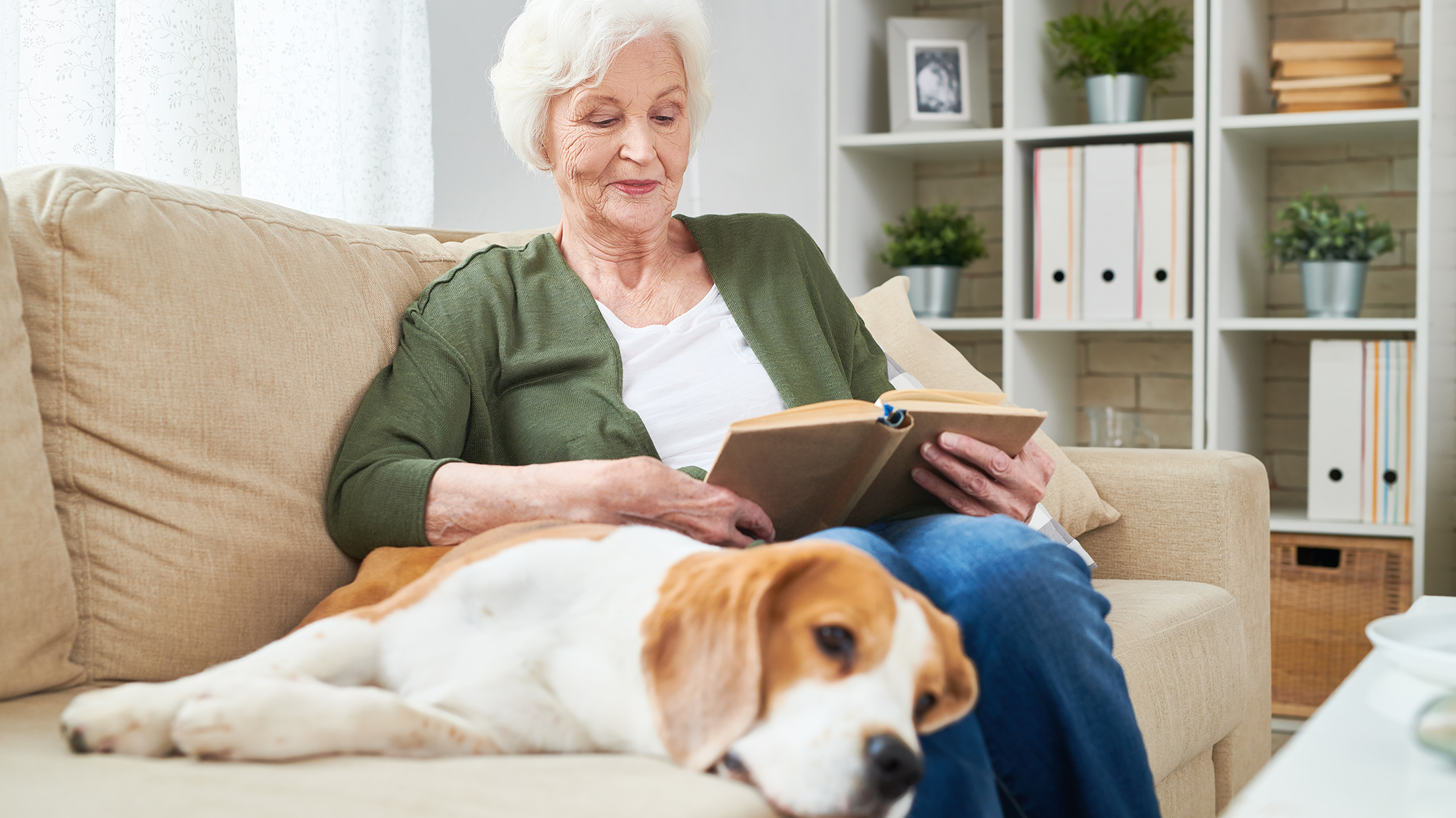 Mujer mayor leyendo un libro en el sofá junto a un perro.